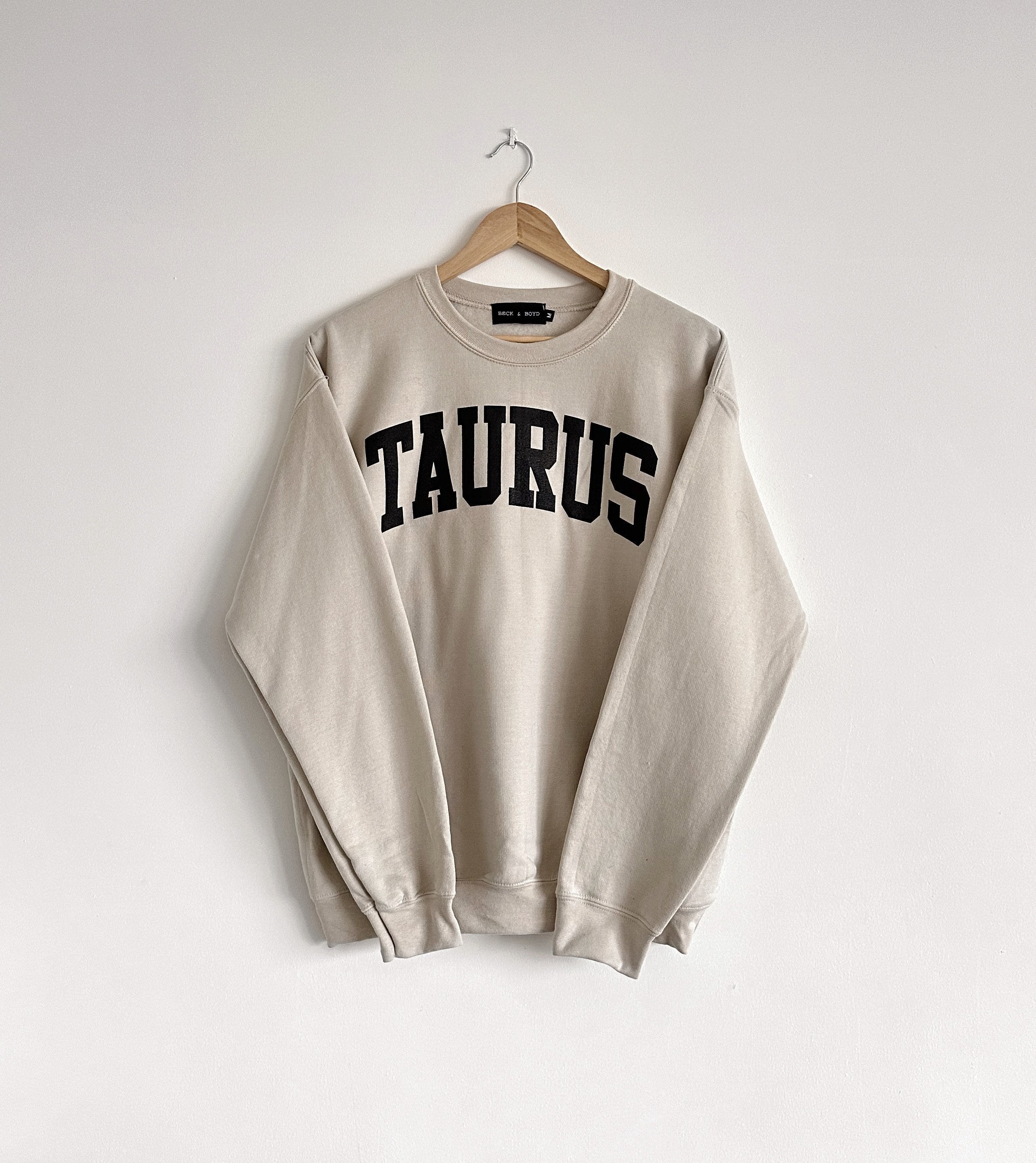 Beige sweatshirt with 'TAURUS' text on a hanger against a white background, Taurus, Zodiac, Astrology, Cozy