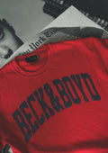 Red sweatshirt with 'BECK & BOYD' text on a surface with a newspaper underneath. Red, Bold, Statement clothing, cozy, IT Girl, bold sweater