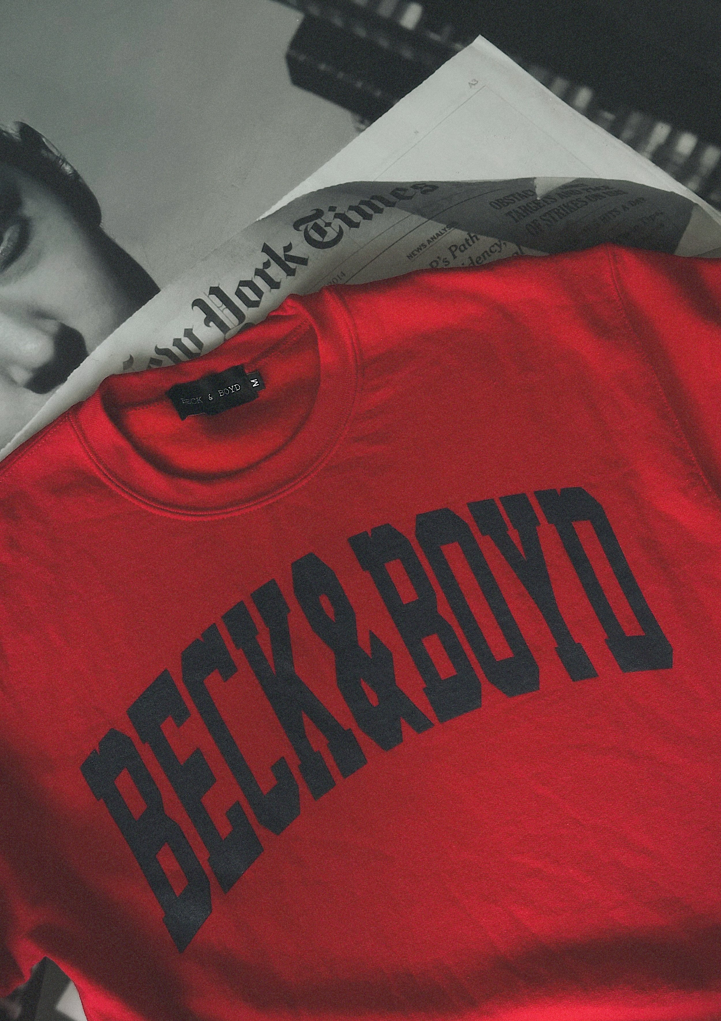 Red sweatshirt with 'BECK & BOYD' text on a surface with a newspaper underneath. Red, Bold, Statement clothing, cozy, IT Girl, bold sweater