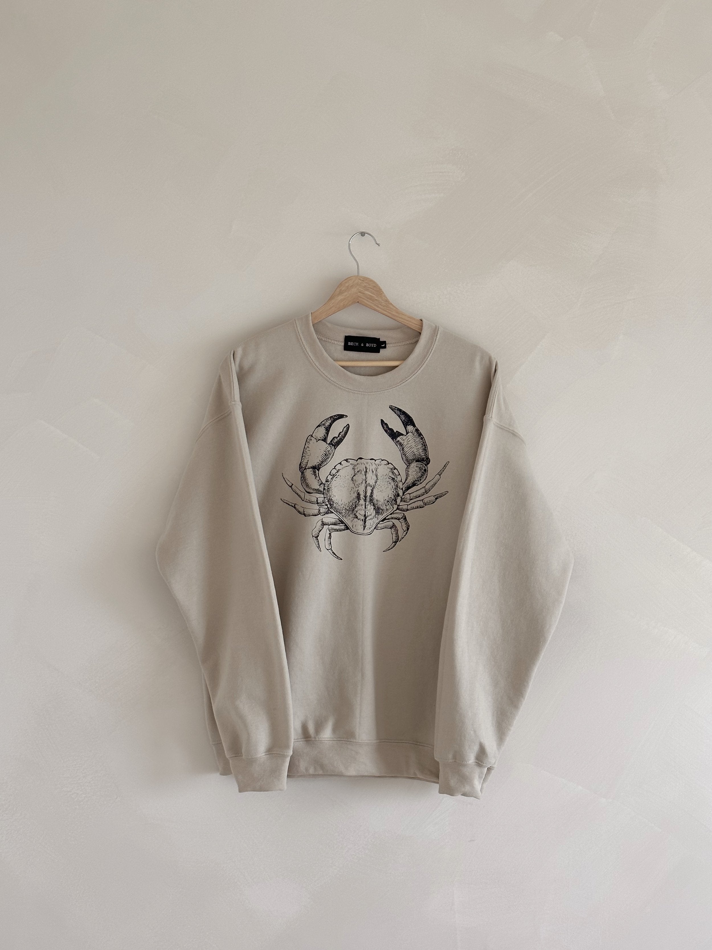 Beige sweatshirt with a crab design on a hanger against a white wall, cancer zodiac, zodiac, trendy