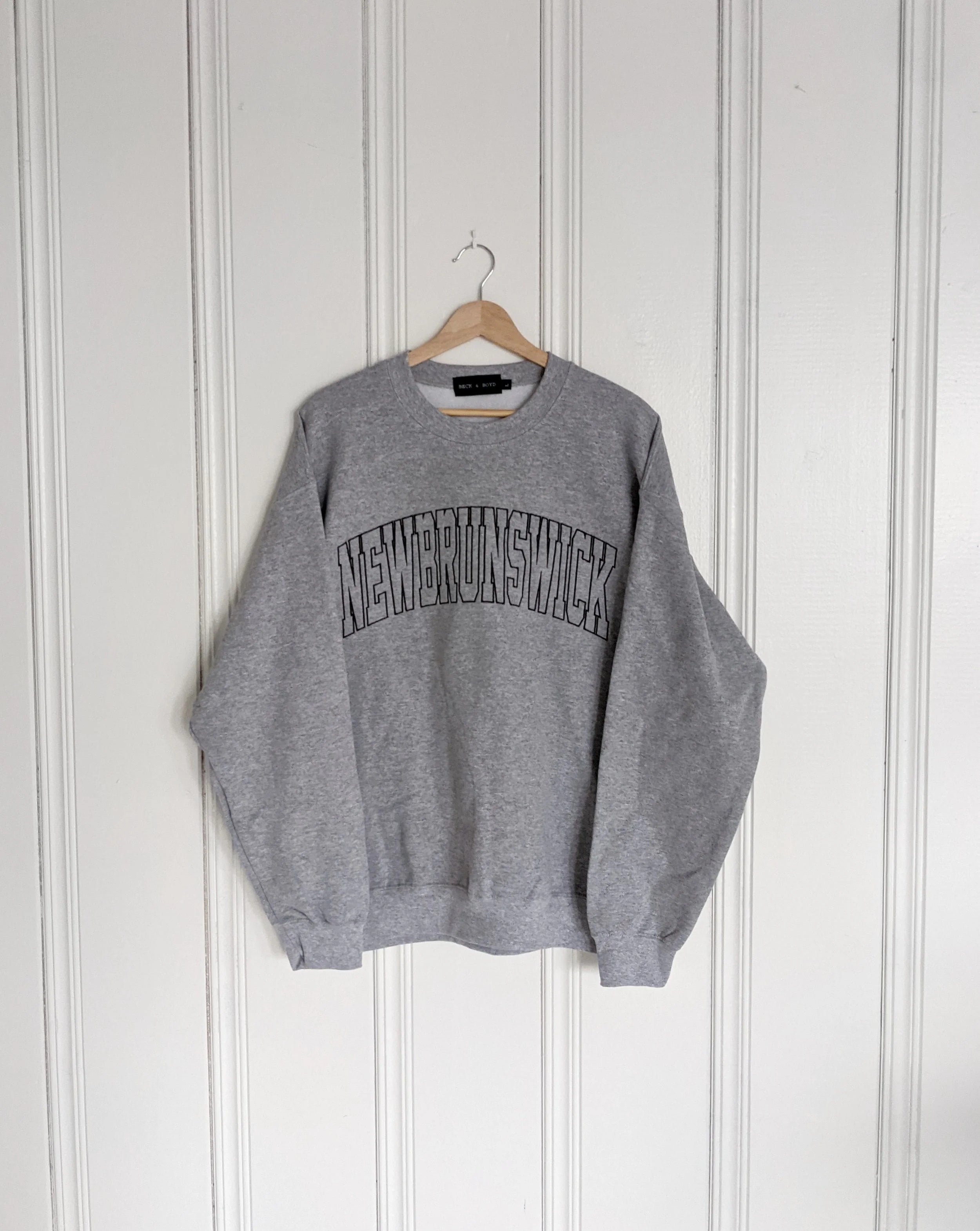 Grey sweatshirt with 'New Brunswick' text on a hanger against a white wall, Embroidered, Cozy, New Brunswick, NB, Maritimes, Gift Ideas