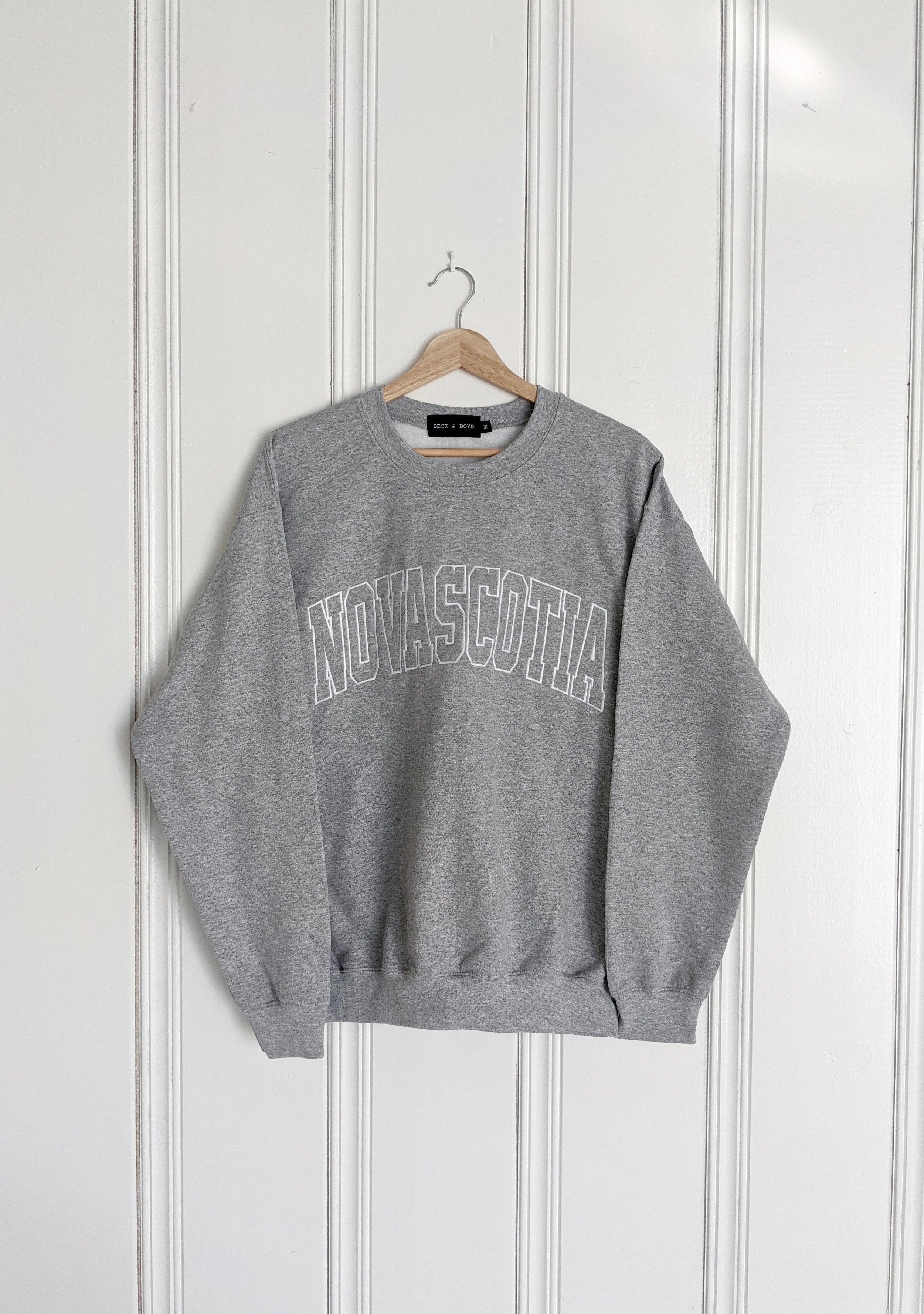 Grey sweatshirt with 'Nova Scotia' text on a hanger against a white paneled wall, Nova Scotia, Cozy, Maritimes, NS