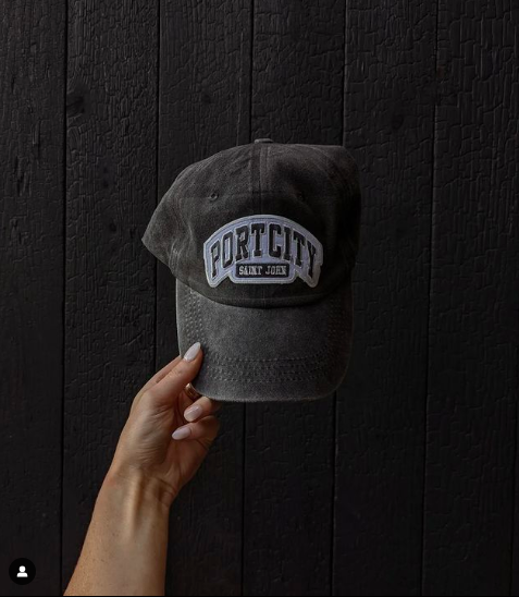 Grey cap with 'PortCity Saint John' logo held against a dark wooden background, ball cap, hat, dad hat, vintage vibe, weathered look, Port City, Saint John
